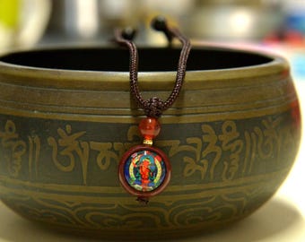 May include: A brown cord necklace with a circular pendant featuring a colorful design and a red bead. The necklace is displayed in front of a bronze-colored bowl with etched patterns and symbols.