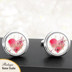 May include: A pair of silver cufflinks with a white background and a red heart with gold lettering that says "XOXO S&J". The heart is surrounded by gold glitter.