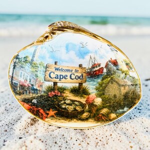 Może przedstawiać: Ozdobna muszla ze złoconym brzegiem, przedstawiająca nadruk z nadmorskim krajobrazem Cape Cod. Obraz ukazuje drewniany znak z napisem Welcome to Cape Cod, czerwoną łódź rybacką, latarnię morską, budynki oraz kosz z ostrygami.
