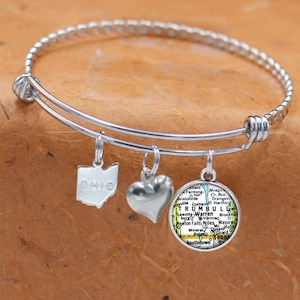 Op de afbeelding: Zilverkleurige armband met drie bedels. Bedels zijn de staat Ohio, een hart en een ronde bedel met een kaart van Trumbull County, Ohio. De armband ligt op een bruin oppervlak.