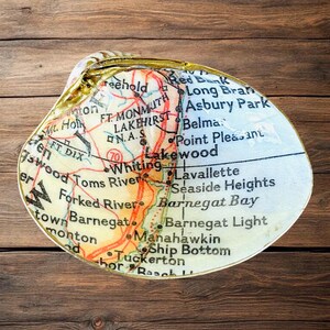 May include: A decorative clam shell dish featuring a printed map of the New Jersey coastline, including locations such as Lakewood, Toms River, and Barnegat Bay. The shell has a smooth, glossy finish with a metallic gold-painted edge along the rim.