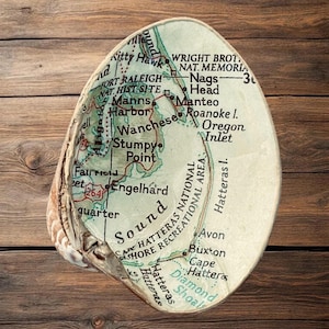May include: A large seashell with a map of the Outer Banks, North Carolina, decoupaged onto its interior surface. The map features text including Wright Brothers National Memorial, Nags Head, Manteo, Wanchese, Stumpy Point, and Cape Hatteras.