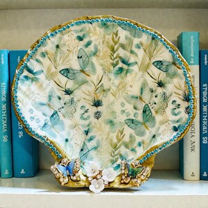 May include: A decorative seashell-shaped tray with a gold rim and a butterfly and floral design. The tray is adorned with blue gemstones around the edge and features a base with white flowers and additional butterfly accents.