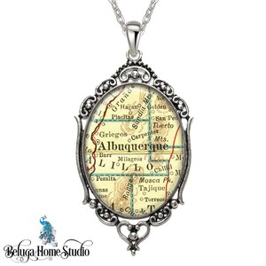 May include: A silver oval pendant necklace featuring a vintage map of Albuquerque, New Mexico. The map shows surrounding towns and landmarks, including Sandia Mountains, Golden, and Placitas.