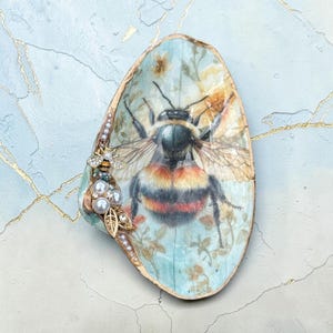 May include: Oval-shaped decorative shell with a painted bumblebee design. The shell features a light blue background with floral accents and a gold-painted rim. Embellished with pearls, a rhinestone bee, and gold-colored leaves.