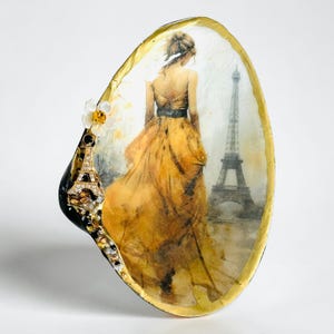 Puede incluir: Arte decorativo en concha marina con una mujer en vestido amarillo y la Torre Eiffel. La concha tiene un borde dorado y un pequeño dije de la Torre Eiffel con una flor. Tema parisino.