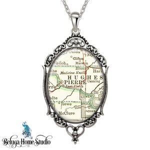 May include: A silver pendant necklace featuring a vintage map of Hughes County, South Dakota. The map shows the towns of Pierre, East Pierre, Canning, and others. The pendant is framed in an ornate silver frame.