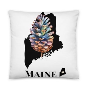 May include: A white throw pillow with a black outline of the state of Maine. A colorful pine cone is centered in the outline. The words "MAINE" are printed below the outline. A small heart is in the upper right corner of the outline.
