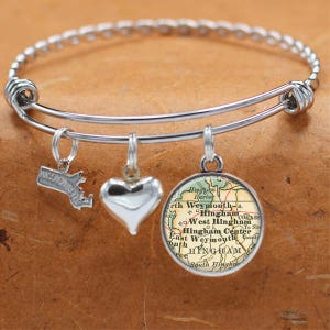 May include: A silver charm bracelet with three charms: a heart, a map of Hingham, Massachusetts, and the state of Massachusetts. The map charm features the text "Hingham Harbor", "North Weymouth", "West Hingham", "Hingham Center", "East Weymouth", and "South Hingham".