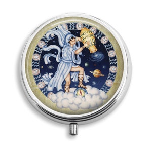 May include: A round, silver pill case with a detailed illustration of a figure pouring water from a golden urn. The artwork features a celestial background with planets, stars, and clouds. The case has a push-button opening.