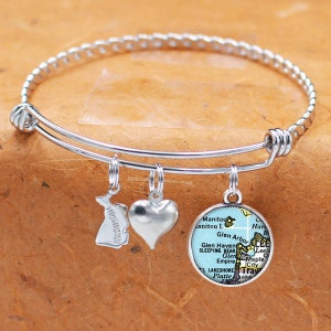 Puede incluir: Una pulsera de brazalete extensible en tono plateado con tres dijes colgantes: una forma recortada del estado de Michigan, un corazón plateado liso y un colgante circular que muestra un mapa impreso de la zona de Glen Arbor y Sleeping Bear.