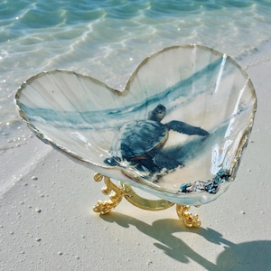 May include: Heart-shaped seashell bowl with a painted sea turtle scene, resting on a gold-colored metal stand. The bowl features blue and white hues, evoking a coastal theme. The bowl is displayed on a sandy beach.
