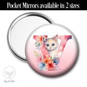 May include: A round pocket mirror with a white background and a pink watercolor letter V with a white kitten peeking out from behind it. The letter V is surrounded by pink, orange, and blue watercolor flowers.