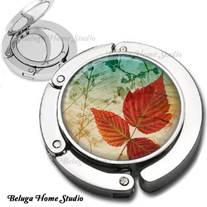 May include: A silver-toned, foldable, round purse hook with a colorful image of autumn leaves on a cream background. The image features two red and orange leaves with a green and blue background.
