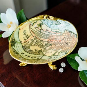 May include: A decorative shell dish featuring a vintage map of Charleston, South Carolina, printed on the interior. The shell has a gold-painted rim and is supported by three small, ornate gold-toned metal feet, creating a raised display bowl.