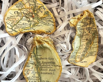 May include: Three oyster shells decorated with sections of vintage maps, including Charleston, St. Augustine, and Cape May. The edges are painted gold, and the shells rest on white paper strips. A unique coastal decor item.