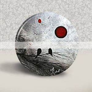 Puede incluir: Una impresión redonda sobre lienzo, de color gris y blanco, con una rama de árbol negra en la que están posados dos pájaros negros. Un globo rojo flota en el cielo sobre la rama del árbol. Un círculo rojo se encuentra en la esquina superior derecha de la impresión.