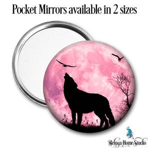 Puede incluir: Una luna llena rosa con un lobo aullando hacia ella. El lobo es una silueta negra contra la luna rosa. Dos pájaros negros vuelan en el cielo. La imagen está en un espejo de bolsillo redondo.