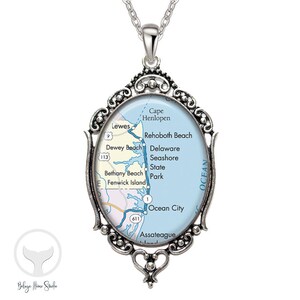 May include: A silver necklace with an oval pendant featuring a map of the Delaware coast. The map shows the towns of Cape Henlopen, Lewes, Rehoboth Beach, Dewey Beach, Bethany Beach, Fenwick Island, Ocean City, and Assateague Island. The pendant is surrounded by a decorative silver frame.