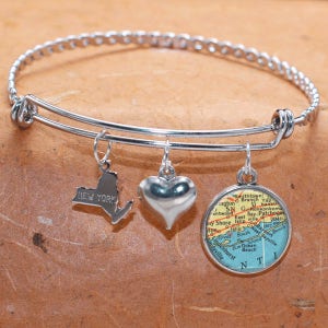 Puede incluir: Un brazalete de plata con tres dijes: un corazón, un mapa de Long Island, Nueva York, y el contorno del estado de Nueva York. El brazalete está hecho de metal plateado y tiene un cierre de resorte.