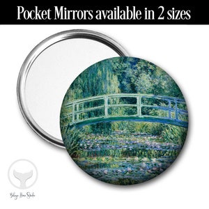 May include: A round pocket mirror with a silver frame and a colorful image of a bridge over a pond with water lilies. The image is a painting of a landscape with green trees and blue water.