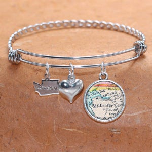 May include: A silver charm bracelet with three charms: a heart, a state of Washington outline, and a vintage map of South Bend, Washington. The bracelet is made of stainless steel and has a braided design.