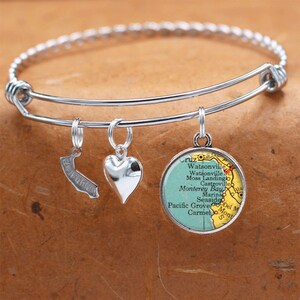 May include: A silver bangle bracelet with three charms: a heart, a California outline, and a round charm with a vintage map of the Monterey Bay area. The map features the names of towns and cities, including Watsonville, Moss Landing, Castroville, Monterey Bay, Marina, Seaside, Pacific Grove, and Carmel.