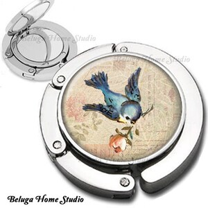 May include: A silver folding compact mirror with a blue bird design on a vintage floral background. The mirror has a hinged lid and a small handle.