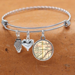 May include: A silver charm bracelet with three charms: a heart, a state of Illinois outline, and a vintage map of a region in Illinois. The map charm has the words "Addison", "Elmhurst", "Glenellyn", "heaton", "AGE", "Lagrange", "Austin", "Allersburg", "rove", "Marchdon", "Hinsdale", and "ALTON" printed on it.