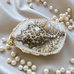 Könnte beinhalten: Eine dekorative Muschel mit Leopardenmuster, goldfarben eingefasst und mit Perlen verziert. Die Muschel liegt auf einer satinartigen Oberfläche, umgeben von verstreuten Perlen, was eine elegante und künstlerische Komposition ergibt.