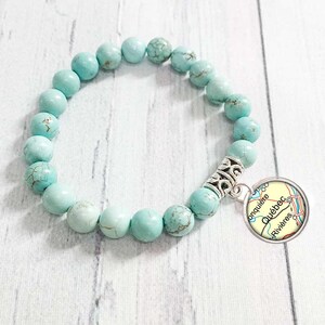 May include: A turquoise beaded bracelet with a silver charm. The charm has a map design with the text "Québec Rivières". The bracelet is made of round, light blue beads.