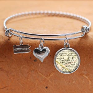 Puede incluir: Un brazalete expandible de plata con tres dijes: un corazón, un contorno del estado de Pensilvania y un dije redondo con un mapa vintage de Doylestown, Pensilvania. El mapa muestra las ciudades circundantes y el texto "BUC" para el condado de Bucks.