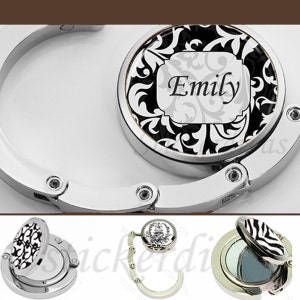 May include: A silver-toned purse hook with a black and white floral design and the name "Emily" in a white rectangle. The hook is closed and ready to be used.