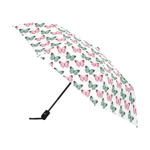 May include: A white umbrella with a repeating pattern of pink and teal butterflies. The umbrella has a black handle and a black button to open and close it. The butterflies are evenly spaced across the surface.
