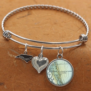 Puede incluir: Un brazalete de plata con tres dijes: un coraz&oacute;n, un mapa de las Outer Banks de Carolina del Norte y un contorno del estado de Carolina del Norte. El dije del mapa tiene el texto "Currituck Light Corolla".