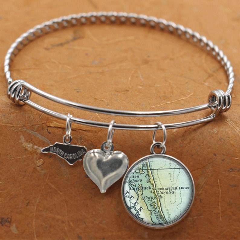 Puede incluir: Un brazalete de plata con tres dijes: un coraz&oacute;n, un mapa de las Outer Banks de Carolina del Norte y un contorno del estado de Carolina del Norte. El dije del mapa tiene el texto "Currituck Light Corolla".