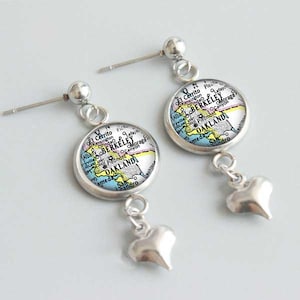 May include: A pair of silver earrings with a heart charm. Each earring features a round, silver-rimmed charm with a vintage map of Oakland, California. The map is in shades of blue, pink, yellow, and green.
