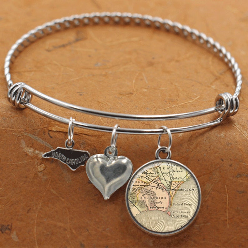 May include: A silver expandable bangle bracelet with three charms: a heart, a map of Wilmington, North Carolina, and an outline of the state of North Carolina. The map charm features the text "BRUNSWICK" and "WILMINGTON".