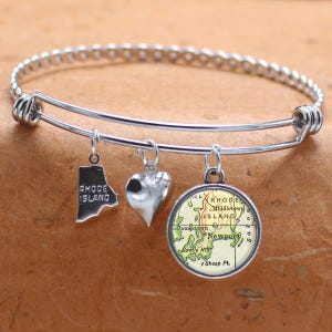 Puede incluir: Un brazalete de plata con tres dijes: un corazón, un contorno del estado de Rhode Island y un mapa de Rhode Island con el texto "Rhode Island" y "Newport".