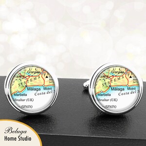 May include: A pair of silver cufflinks with a round, clear glass design. The glass features a map of the Costa del Sol region of Spain, with the cities of Malaga, Motril, and Marbella labeled. The text "SISTEMA" is also visible on the map.