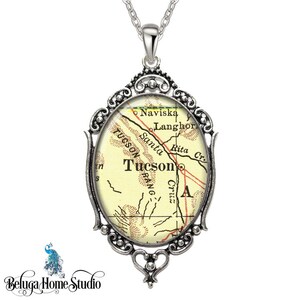 May include: A silver pendant necklace with an oval-shaped glass charm featuring a vintage map of Tucson, Arizona. The map shows the city's streets and landmarks, including the Santa Cruz River and the Tucson Mountains.