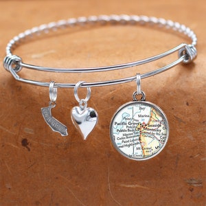 May include: A silver charm bracelet with three charms: a heart, a California outline, and a round charm with a map of the Pacific Grove area of California. The map shows the cities of Pacific Grove, Seaside, Monterey, and Carmel.