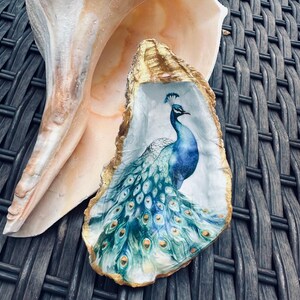 May include: A decorative oyster shell featuring a painted peacock design. The peacock is depicted in shades of blue and green, with a detailed tail. The shell has a gold-painted edge and rests against a larger, natural shell.