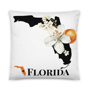 May include: A white pillow with a black outline of the state of Florida, with white flowers and an orange fruit. The text "FLORIDA" is printed in black below the outline. A smaller outline of Florida with a black heart is in the bottom left corner.