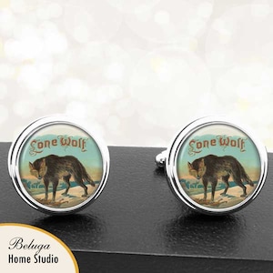 May include: A pair of silver cufflinks with a vintage illustration of a wolf on a blue background. The cufflinks feature the text "Lone Wolf" in a decorative font.