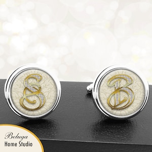Puede incluir: Un par de gemelos de plata con fondo blanco y letras doradas. Las letras son "S" y "B" y están decoradas con pequeños diamantes de imitación blancos.