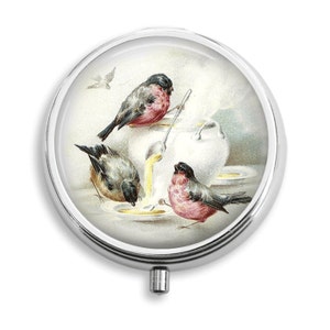 May include: A round, silver-colored pill box with a hinged lid. The lid features a vintage illustration of three birds, two gray and one pink, drinking from a white bowl. The bowl is filled with a yellow liquid.