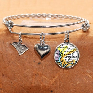 May include: A silver bangle bracelet with three charms: a heart, a state of South Carolina outline, and a map of South Carolina with the text "Clemson, Pendleton, La France, Sandy Springs".