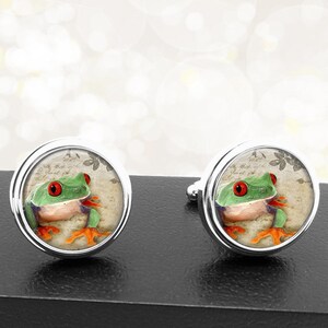 May include: A pair of silver cufflinks with a round, glass-like design featuring a green tree frog with red eyes on a vintage-style background.