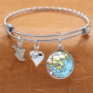 Może przedstawiać: Srebrna bransoletka z trzema zawieszkami: sercem, mapą Maryland i mapą Dundalk w Maryland. Zawieszki z mapami są w stylu vintage.